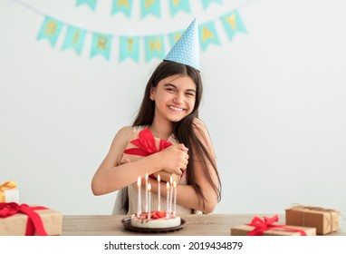 Happy Indian teen girl in party hat sitting at table, hugging gift box, having birthday cake with candles at home. Cheerful Asian adolescent celebrating holiday alone, looking at camera