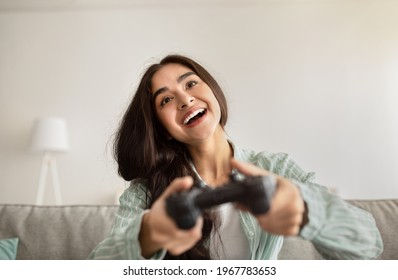 Happy Indian Woman With Joystick Playing Online Video Games, Laughing And Having Fun At Home. Positive Young Lady With Controller Enjoying Computer Gaming. Weekend Activities Concept