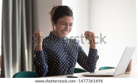 Happy indian woman office worker feeling excitement raising fists celebrates career ladder promotion or reward, businesswoman sitting at desk receive online news, great results successful work concept