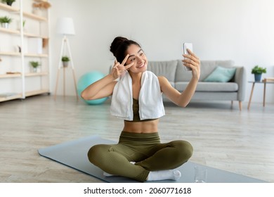 Happy Indian Woman Taking Selfie During Home Workout, Sitting On Yoga Mat Indoors, Full Length. Millennial Asian Lady With Cellphone Making Photo Of Herself On Domestic Training