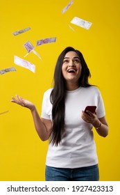 Happy Indian Woman Using Smartphone And Flying Or Raining Or Falling Money, Rupee Banknotes On Yellow Color Studio Background