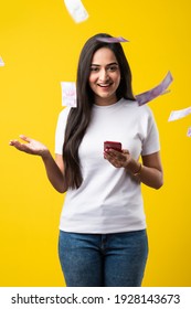 Happy Indian Woman Using Smartphone And Flying Or Raining Or Falling Money, Rupee Banknotes On Yellow Color Studio Background