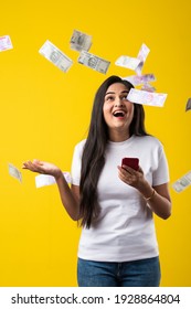 Happy Indian Woman Using Smartphone And Flying Or Raining Or Falling Money, Rupee Banknotes On Yellow Color Studio Background