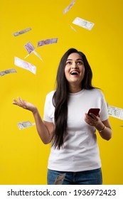 Happy Indian Woman Using Smartphone And Flying Or Raining Or Falling Money, Rupee Banknotes On Yellow Color Studio Background