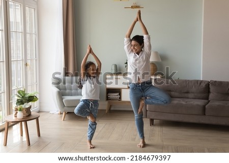 Happy Indian yogi mom teaching daughter kid to do yoga at home, standing in tree pose, smiling, laughing, talking, enjoying home exercises, leisure activities, keeping healthy life style