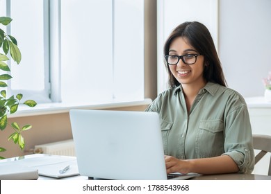 Happy indian young adult woman wearing glasses using pc laptop computer working studying at home office sitting at table. Happy female professional freelancer learning watching online webinar training - Shutterstock ID 1788543968