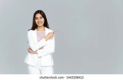 Happy Indian Young Business Woman Wear White Suit Looking At Camera Pointing Finger At Copy Space Isolated On Grey Studio Background, Smiling Beautiful Confident Hindu Lady Professional Showing Aside