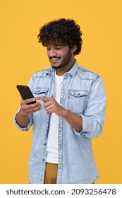 Happy indian young man using cell phone isolated on yellow background. Smiling ethnic hipster guy holding smartphone playing game in app, dating buying online in ecommerce store on cellphone. Vertical
