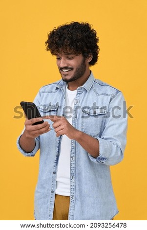 Happy indian young man using cell phone isolated on yellow background. Smiling ethnic hipster guy holding smartphone playing game in app, dating buying online in ecommerce store on cellphone. Vertical