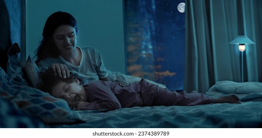 Happy Indian young mother with little daughter child lying on bed night bedtime at indoor house bedroom. beautiful mummy stroking hand on small girl head sleeping nighttime dark home - Powered by Shutterstock