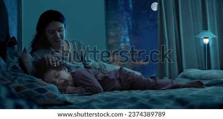 Happy Indian young mother with little daughter child lying on bed night bedtime at indoor house bedroom. beautiful mummy stroking hand on small girl head sleeping nighttime dark home