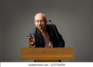 Happy Indian/asian Handsome Senior Businessman Speaking With Mic At Podium In Office Or Auditorium Presenting Something
