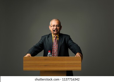 Happy Indian/asian Handsome Senior Businessman Speaking With Mic At Podium In Office Or Auditorium Presenting Something
