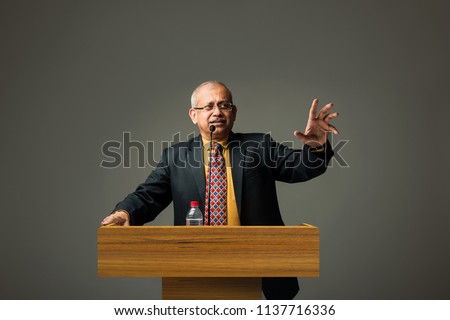 Happy Indian/asian handsome senior businessman speaking with mic at podium in office or auditorium presenting something
