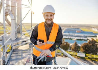 A happy industry worker holding phone on height on metal construction and smiling.