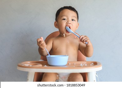 Happy Infant Asian Baby Boy Sitting On Baby High Chair And Eating Food By Himself And Making Mess.