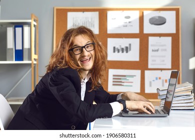 Happy Insane Male Office Worker In Glasses Sitting At Table, Working On Laptop Computer, Smiling And Looking At Camera. Funny Crazy Employee Or Accountant With Long Messy Hair Doing Financial Reports