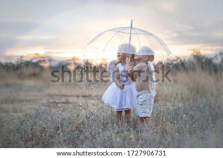 Happy and inspired angels boy and girl stand in the pouring rain hiding under an umbrella. A rainbow is visible in the sky. Thin streams of rain are barely visible on the whole background. 