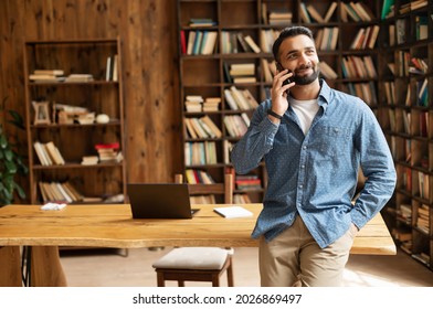 Happy inspired Indian male small business owner talks on the smartphone standing in modern loft office, smiling successful contemporary man manager in casual wear holding phone call, copy space