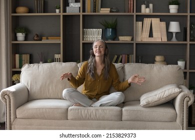 Happy Inspired Mature Lady In Wireless Earphones Meditating To Lounge Relaxing Music, Sitting In Lotus Pose On Cozy Couch, Making Dance Hands, Smiling With Closed Eyes, Practicing Soft Yoga