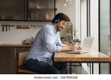 Happy Interested Millennial Generation Man In Headphones Listening Educational Lecture, Studying On Online Courses In Modern Kitchen, Handwriting Notes In Paper Copybook, Improving Knowledge.