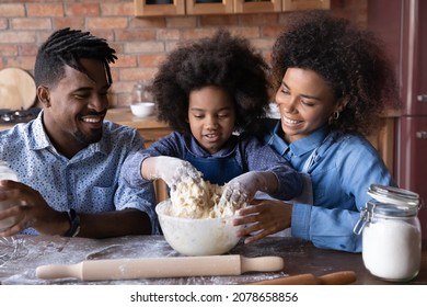 Happy Interested Small Cute Child Girl Preparing Dough For Homemade Pastry With Joyful Caring Young African American Couple Parents, Having Fun Cooking Baking Together In Kitchen On Weekend