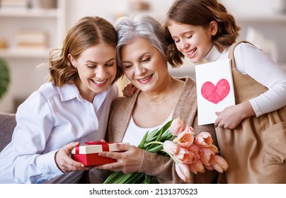 Happy International Women's Day. Multi Generational Family Daughter And Granddaughter Giving Flowers  And Gift To Grandmother  Cheerfully Celebrate The Spring Holiday Mother's Day Or Grandparents Day 