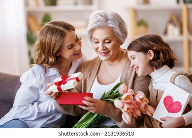 Happy International Women's Day.  Multi Generational Family Daughter And Granddaughter Giving Flowers  And Gift To Grandmother  Cheerfully Celebrate The Spring Holiday Mother's Day Or Grandparents Day