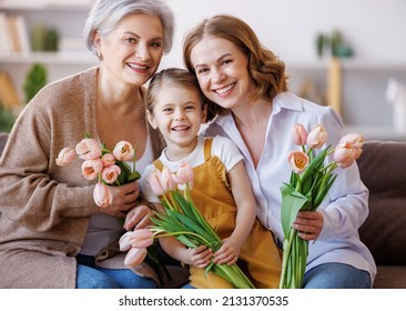 Happy International Women's Day. Smiling Multi Generational Family Grandmother, Mother And Daughter With Flowers  Cheerfully Celebrate The Spring Holiday Mother's Day At Home