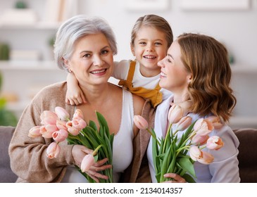 Happy International Women's Day. Smiling Multi Generational Family Grandmother, Mother And Daughter With Flowers  Cheerfully Celebrate The Spring Holiday Mother's Day At Home