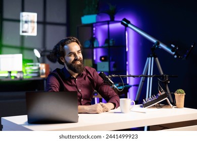 Happy internet star using quality mic in studio to discussing with viewers. Smiling man sitting at table in front of laptop using high tech recording gear to record social media video