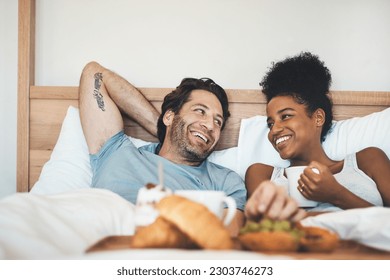 Happy interracial couple, bed and breakfast in relax for morning, bonding or relationship at home. Man and woman smiling with food, coffee or meal relaxing on holiday or weekend together in bedroom - Powered by Shutterstock