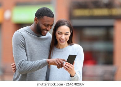Happy interracial couple checking cell phone laughing outdoors in the street