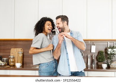 Happy interracial couple dancing in the kitchen, singing while cooking breakfast or dinner, young African American woman and a handsome man smiling and having fun together, holding kitchen utensils - Powered by Shutterstock