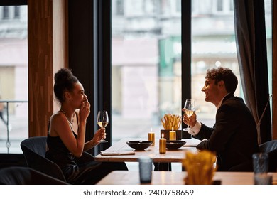 happy interracial couple in elegant attire holding glasses of wine during date in restaurant, laugh - Powered by Shutterstock