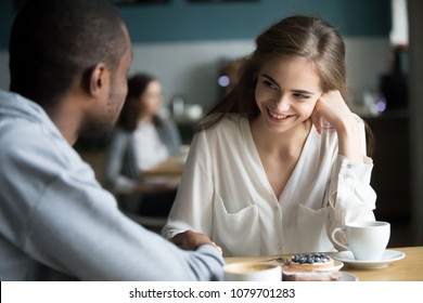 Happy interracial couple flirting talking sitting at cafe table, african man holding hand of smiling caucasian woman having fun drinking coffee together at meeting, biracial lovers on date concept