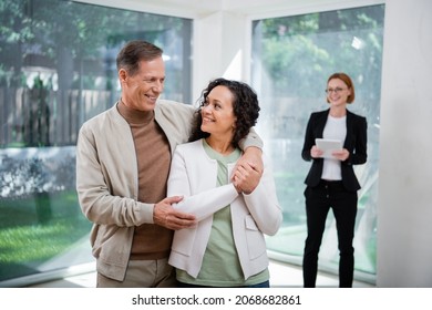 Happy Interracial Couple Hugging Near Redhead Realtor In Glasses Holding Digital Tablet On Blurred Background