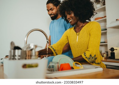 A happy interracial couple is preparing dinner and washing dishes at home together. A happy woman is washing plates and doing dishes in the kitchen sink while her husband is cooking lunch next to her. - Powered by Shutterstock