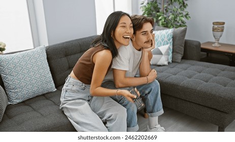 A happy interracial couple sitting together on a gray sofa at home, with the woman laughing and man holding a remote. - Powered by Shutterstock