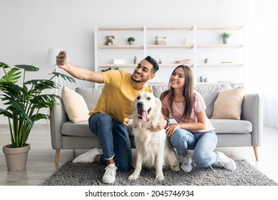 Happy Interracial Couple Taking Selfie With Their Dog While Sitting On Soft Carpet At Home, Full Length. Positive Arab Guy And His Girlfriend Making Photo With Cute Pet In Living Room