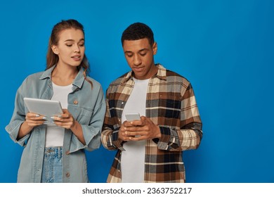 happy interracial couple using  smartphone and digital tablet while standing on blue backdrop - Shutterstock ID 2363752217