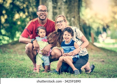 Happy Interracial Family Is Enjoying A Day In The Park. Little Baby Girl And Boy. Successful Adoption. Diverse Family In Nature With Sun In The Back.