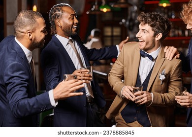 happy interracial friends in formal wear congratulating groom in bar, men holding glasses of whiskey