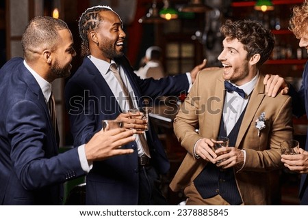 happy interracial friends in formal wear congratulating groom in bar, men holding glasses of whiskey