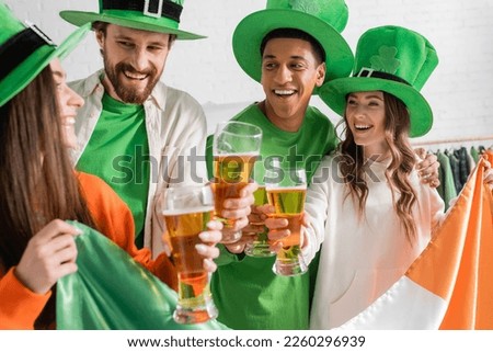 happy and interracial friends in green hats holding glasses of beer and Irish flag while celebrating Saint Patrick Day 