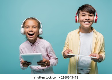 Happy interracial pupils in headphones holding digital tablets isolated on blue - Powered by Shutterstock