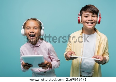 Happy interracial pupils in headphones holding digital tablets isolated on blue