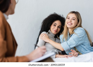 Happy Interracial Same Sex Couple Embracing Near Blurred Psychologist Writing On Clipboard