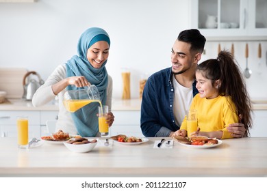 Happy Islamic Family With Little Daughter Having Tasty Lunch In Kitchen Together, Cheerful Middle Eastern Parents And Cute Female Child Eating Delicious Food And Drinking Orange Juice At Home