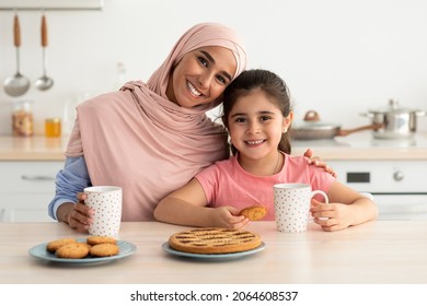 Happy Islamic Family Mom And Little Daughter Eating Snacks In Kitchen Together, Portrait Of Young Middle Eastern Lady In Hijab And Her Cute Female Child Enjoying Homemade Cookies And Drinking Tea